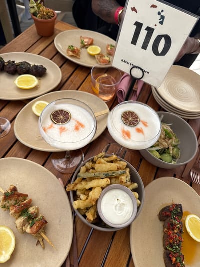 The image shows an outdoor dining setting at Kassi Beach House in Marina del Rey, California. A wooden table is laden with several plates of food and drinks. In the foreground, there are two skewers of grilled meat, one with a garnish of red chili and herbs, and another with chunks of chicken and parsley. A small bowl of white dip sits between them, next to a bowl of fried vegetables. A cocktail with a foamy top and a dried lime garnish is also visible, along with a glass of amber-colored liquid and a small salad. In the background, a table number "110" is propped up on a stand, with the text "KASSI VENICE BEACH" below it. A small potted succulent sits on the edge of the table. The overall scene suggests a relaxed and enjoyable meal outdoors.