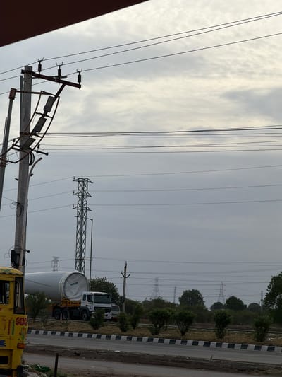 The image depicts a truck carrying a large wind turbine blade on a road in Sugalimetta, India. The truck, a white Mercedes-Benz Actros, is positioned on the right side of the frame, with the massive white blade, branded "SUZLON," dominating the center. A yellow vehicle is partially visible on the left. The scene is set against a backdrop of a cloudy sky and features several power transmission towers and poles, indicative of an industrial or utility area. Green trees and bushes line the roadside, with a checkered concrete barrier separating the road from the land beyond. The overall impression is one of transportation and infrastructure development.