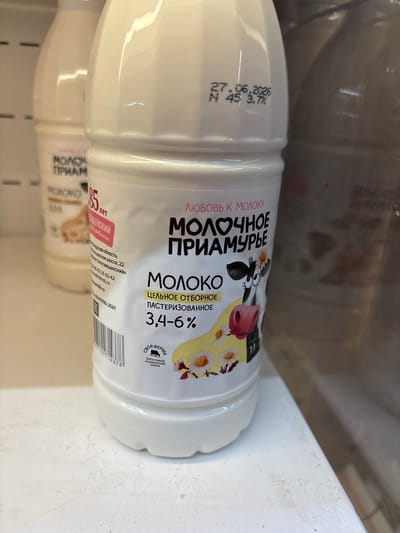 A close-up, eye-level shot shows a white plastic bottle of milk with Russian labeling. The bottle is partially obscured by another similar bottle in the background, also out of focus. The label features a cartoon cow with daisies, and text including "МОЛОЧНОЕ ПРИАМУРЬЕ" (Molochonoye Priamurye), which translates to "Priamurye Dairy." A date, "27.06.2026," is stamped on the bottle. The lighting is bright, suggesting an indoor setting like a store or refrigerator. No people are visible, and the environment beyond the bottles is indistinct.