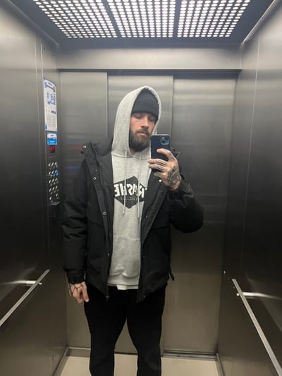 A man is taking a selfie in an elevator. He is wearing a beanie, a gray hoodie, and a black jacket, with black pants. His hands are tattooed. The elevator has a panel of buttons and an illuminated wall. The location cannot be determined based solely on the image.