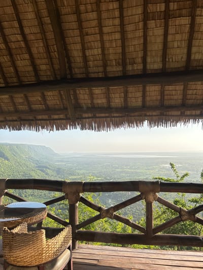 The image captures a scenic overview from an elevated vantage point, likely a lodge or resort in Mto Wa Mbu, Tanzania. The perspective looks out over a vast, densely forested valley or mountainous landscape, extending towards a hazy horizon under a bright daytime sky. In the far distance, a significant body of water, possibly a lake or large river, is visible, partially obscured by atmospheric haze.
In the upper left quadrant of the frame, the underside of a rustic thatched roof structure is prominently featured. The roof is composed of tightly woven natural materials supported by substantial dark wooden beams, showcasing traditional craftsmanship. The uneven, fringed edge of the thatch runs horizontally across the upper middle portion of the image, defining the sheltered area.
To the right, a sturdy wooden balcony railing provides a natural frame for the expansive view. The railing is crafted from thick, dark timber posts and crossbeams, robustly secured at their joints with intricate wrappings of natural cord or rope. In the bottom right corner, a portion of a woven, natural-fiber chair and a small, round table are visible, suggesting a tranquil spot for relaxation and observation.
The scene conveys a sense of calm and natural beauty, characteristic of an ecological retreat. The lighting indicates it is daytime, with a soft, diffused light possibly due to the atmospheric haze over the distant landscape. No text is visible in the image.