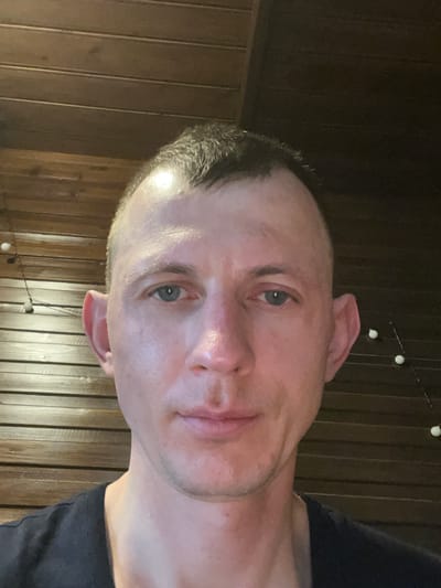A medium shot portrait of a fair-skinned man with short, dark hair and blue eyes is captured from the chest up. He is looking directly at the camera. The man has large ears that stick out. His skin appears slightly oily, and there are some faint blemishes on his face. He is wearing a dark t-shirt. The background consists of a wooden paneled wall. To the left and right of the man, strings of small, round, white lights are visible against the wooden wall, suggesting an indoor or outdoor setting at dusk or night. The lighting appears to be artificial, coming from above and slightly in front of the subject. The scene is devoid of any other people or significant objects. This image does not provide any specific geographic or locational information that could pinpoint it to Zakopane, Poland.