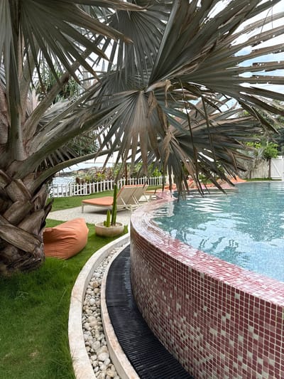 A serene outdoor setting features a palm tree in the foreground. Adjacent to the palm is a curvilinear pool with red mosaic tiles. Water cascades from the pool's edge into a curved basin filled with white stones, surrounded by manicured grass. In the background, white chairs are partially visible, alongside a white fence. The scene suggests a luxurious or resort-like atmosphere.