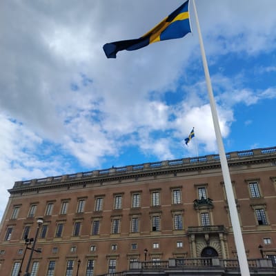 The Royal Palace in Stockholm, Sweden, is captured in a low-angle photograph under a partially cloudy sky displaying both blue and white-grey tones. The building, constructed with a light brown facade, spans multiple stories and features numerous rectangular windows, many accented with decorative surrounds. A tall, white flagpole is positioned in the mid-left foreground, partially obscuring the palace's structure. From this pole, a large Swedish national flag, characterized by a blue field with a yellow Nordic cross, unfurls horizontally to the left. A smaller, identical Swedish flag flies from a shorter pole located on the building's roofline towards the upper-right. Architectural details visible on the palace include a decorative stone balustrade along the roof and an ornate entrance section on the lower levels, featuring a balcony supported by columns above an arched portal. A small, dark human figure is discernable standing on the rooftop, behind the balustrade, to the left of the smaller flag. A black street lamp is partially visible on the far-left edge of the frame, and a stone balustrade forms the lower boundary of the foreground.