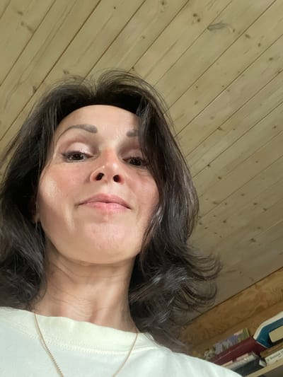 An over-the-shoulder, low-angle, close-up shot shows a middle-aged woman from the chest up, looking directly at the camera. She has dark brown hair that frames her face and falls past her shoulders. Her skin is fair with a slight tan. She is wearing light-colored clothing, possibly a white or cream-colored t-shirt, with a delicate gold necklace visible. The background is a wooden paneled wall, characteristic of an interior space. The image does not provide enough information to determine a specific location within the city of Kumyony, Russia, as the setting is generic.