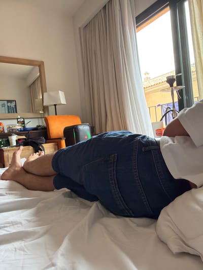 A medium shot depicts a person wearing a white shirt and denim shorts, positioned on a white bed next to a window. The window appears to be a skylight type, revealing a view of buildings outside. Adjacent to the bed is a shelf or nightstand with various items including bottles, a black speaker, and an orange chair. The room also features a mirror reflecting part of the interior. The Burger King packaging hints at fast food consumption. The location is potentially Punta Umbría, Spain, as inferred from the context. The focus is on the individual in the bed, with additional elements providing context to the setting.
