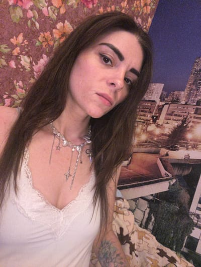 The image shows a close-up, low-angle shot of a young woman with long, dark hair lying down. She is wearing a white top with lace trim and a silver necklace with star and crescent moon charms. Her expression is neutral, with her head tilted slightly. The background appears to be a bedroom with a wallpaper depicting a cityscape and a floral patterned bedding. The lighting suggests it could be indoors during the day, possibly with artificial light. There are visible tattoos on her right arm. The scene does not depict any specific activity or event beyond the subject posing for the photo. The location context provided is Krasnoe-na-Volge, Russia, but there are no explicit visual cues in the image to confirm this. No text is visible in the image.