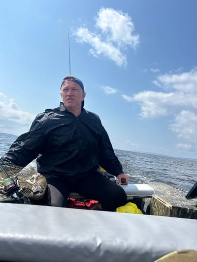 An adult male wearing a blue baseball cap and a black jacket is in a boat on a body of water. He is looking to his right and appears to be talking. A fishing rod is visible behind his head, and a fishing net and reel are in the foreground to his left. The boat is moving, as indicated by the wake in the water behind it. The sky is blue with scattered white clouds. The location appears to be a natural environment, possibly a lake or river, and is consistent with the geography of Kostovaty, Russia.