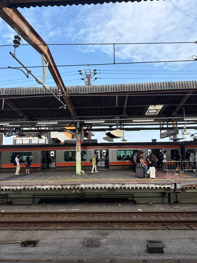 This is a medium-shot, eye-level, outdoor photograph taken at a train station in Funabashi, Japan, under a partly cloudy sky. The scene captures a train at a platform, with passengers boarding and disembarking. The train is orange and gray, with multiple windows and doors. Several individuals are visible on the platform, some with luggage, suggesting travel. The platform itself is marked with yellow and white lines, indicating safety zones. Overhead, a corrugated roof is supported by metal beams and wires, typical of train station infrastructure. In the background, buildings are visible. The tracks and gravel area in the foreground indicate the active railway environment.