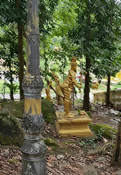 A golden statue of a mythical creature, possibly a Kinnara or Apsara, stands on a golden pedestal in a jungle-like environment. The statue depicts a figure with human-like torso and legs, adorned with elaborate headdress and jewelry, and possessing bird-like legs and tail feathers. It holds a sword-like object in its right hand and its left arm is bent. The statue is surrounded by lush green foliage, moss-covered rocks, and fallen leaves on the ground. In the foreground, a weathered stone pillar with decorative carvings and faded yellow paint stands prominently. Further in the background, hints of a temple or pagoda structure with golden embellishments are visible through the trees. The scene suggests a religious or cultural site within the tropical landscape of Sihanoukville, Cambodia.