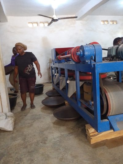 The image depicts an indoor industrial workspace in Olomoda, Nigeria, containing a large processing machine and three individuals. The room features a concrete floor, light-colored walls with some discoloration, and a white ceiling from which a black three-bladed ceiling fan and a bright light fixture are suspended. Two small rectangular openings are visible high on the far wall.
Dominating the mid-ground is a robust industrial machine, primarily constructed with a blue metal frame, featuring red pulleys, black belts, and a red hopper on its top left. A conveyor belt is visible on the right side of the machine. The machine rests on wooden planks, elevating it from the floor. Beneath the machine, arranged on the floor, are five dark, shallow, circular metal pans, seemingly positioned to collect output. An engine or motor assembly is visible on the upper right side of the machine, suggesting mechanical operation.
To the left of the machine, two dark plastic buckets or basins are on the floor. A white sack, partially visible on the far left, bears some obscured English text.
Three individuals are present. In the center-left foreground, a person wearing a light-colored head covering, a dark t-shirt with "Fila" written in white, dark shorts, and light-colored flip-flops, walks towards the right, looking downwards. Their left arm is bent, and the right arm is slightly extended. Partially obscured behind this individual, another person, wearing a purple shirt and light-colored trousers, appears to be carrying a dark, flat object. A third individual is partially visible on the far right, positioned behind the machine, with only their head and upper torso discernible, suggesting engagement with the machinery.