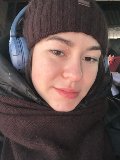 A young adult female subject is depicted in a close-up, selfie-style photograph taken in the city of Pyriatyn, Ukraine. She is wearing a dark brown knit beanie and light blue over-ear headphones. Her attire also includes a dark-colored puffer jacket and a dark brown knit scarf wrapped around her neck, covering the lower portion of her face. She has brown eyes, a slight smile, and is wearing light pink lipstick. The subject is looking directly towards the camera. The background is blurred and indistinct, suggesting an indoor or confined environment.
