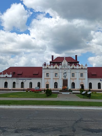 The image captures a frontal view of a two-story train station building with a distinctive red tiled roof, located in the city of Парэчча, Belarus. The building's facade is cream-colored with beige decorative elements around the windows and cornices. A statue of a soldier stands prominently in front of the central section of the building, positioned on a stone pedestal adorned with a wreath of flowers. The area in front of the station is paved with gray bricks and surrounded by green grass. A red car is parked on the left side of the building, and a street lamp is visible between the car and the entrance. The sky above is filled with a mix of white and gray clouds. The scene appears to be an outdoor, public space, likely a train station plaza.