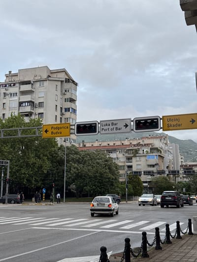 The image shows a street scene in Polje, Montenegro. The view is perpendicular to the street. Road signs indicate directions to Ulcinj/Skadar, Luka Bar/Port of Bar, and Podgorica/Budva. A traffic light is illuminated with a red light. A pedestrian crosswalk is visible. Vehicles, including a white minivan, are present on the street. Buildings can be seen in the background. A row of black metal posts with chains lines the sidewalk. Trees and vegetation are also visible along the street. The sky is overcast. Some people are walking on the street.