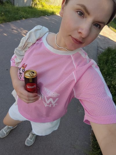 The media file is a selfie taken outdoors, likely during the day with sunny weather, as indicated by the shadows and bright lighting. The subject is a young woman with light skin and blue eyes, wearing a pink mesh jersey over a white top, white shorts, and grey sneakers. She is holding a can of Coca-Cola in her right hand. A light-colored jacket is draped over her left shoulder. She is wearing a pearl necklace. The background shows a paved surface, possibly a road or path, bordered by green grass and a fence. The woman appears to be looking at the camera with a neutral expression. The visible text on the Coca-Cola can reads "Coca-Cola". The image's location context is Korosten, Ukraine.