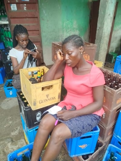 In Yenegoa, Nigeria, two women are seen amidst stacks of beverage crates outside a building with a green, rough-textured wall. The woman in the foreground, wearing a coral-pink dress and a patterned dark blue skirt, sits on a blue plastic crate, leaning her head on her left hand and holding a pink pen in her right. She appears to be looking at a piece of paper. Next to her, a yellow Guinness crate contains several brown bottles, some with yellow labels. Behind her, to the left, a younger woman with braided hair, wearing a white off-the-shoulder top, sits on black plastic crates and looks intently at her smartphone. Several black and blue plastic crates, some filled with bottles, are stacked around the women. The ground is a rough, greyish surface.