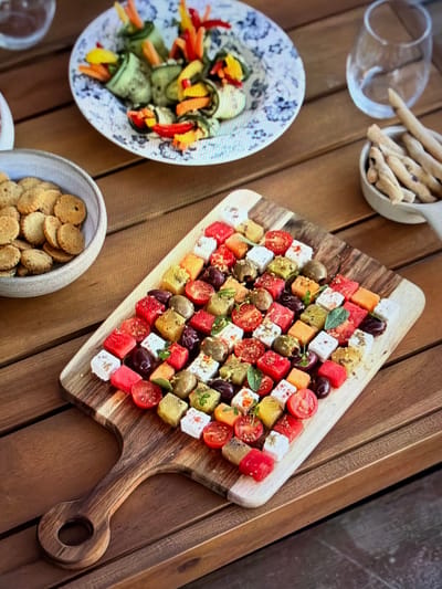 A vibrant outdoor food spread is captured on a slatted wooden table, likely set for a social gathering in Ros Na Rí, Ireland. The central focus is a large, light-colored wooden serving board, shaped with a handle, laden with an array of colorful appetizers. These appear to be individual cubes or small skewers, meticulously arranged in a grid pattern. The cubes showcase a variety of ingredients: bright red pieces (likely watermelon or cherry tomatoes), creamy white feta cheese, dark and light green elements (olives, possibly kiwi or cucumber), and orange/yellow cubes (possibly melon or bell peppers). Small sprigs of fresh herbs, such as mint or thyme, garnish some of the cubes, enhancing their visual appeal.

To the left of the board, a white ceramic bowl with a subtle blue pattern holds neat rolls, possibly made from zucchini or cucumber slices, filled with julienned vegetables in striking red, yellow, and orange. In the top-left, a light-colored bowl is filled with long, thin breadsticks (grissini). Another light-colored bowl at the bottom-left contains a pile of small, round, golden-brown crackers or biscuits. Two clear, empty glasses are partially visible, suggesting beverages are part of the setting. The bright, natural light indicates a daytime event, creating a fresh and inviting atmosphere for an outdoor meal or snack. No text is visible in the image.