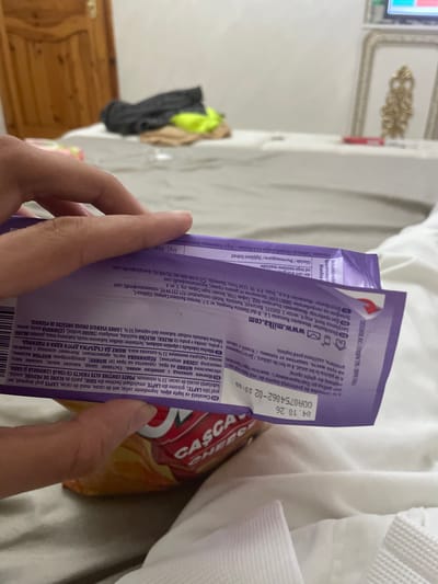 A person's hands are prominently featured in the foreground, holding a purple Milka chocolate bar wrapper. The wrapper displays a best before date of "04.10.26" and a batch code "00A0754062-02 20:00". Text on the packaging, predominantly in Romanian, Bulgarian, and Slovenian, identifies the product as "Alpine milk chocolate" and lists ingredients such as milk, soy, hazelnuts, and wheat, indicating potential allergens. Distributor information for Mondelēz in Romania, Bulgaria, and Slovenia is also visible.

The scene appears to be an indoor setting, likely a bedroom or hotel room in Ihtiman, Bulgaria. In the background, white bed linens or a blanket are partially visible on the right, suggesting a bed. To the left, a portion of a decorative gold and white wall molding is visible, along with a wooden headboard or piece of furniture. Further back, some clothes, including a neon yellow item and a black bag, are draped over an unseen object. Another food item, possibly a bag of chips or crisps, is partially obscured behind the chocolate wrapper. The overall lighting suggests an indoor environment. The person's activity seems to be unwrapping or preparing to eat the chocolate.