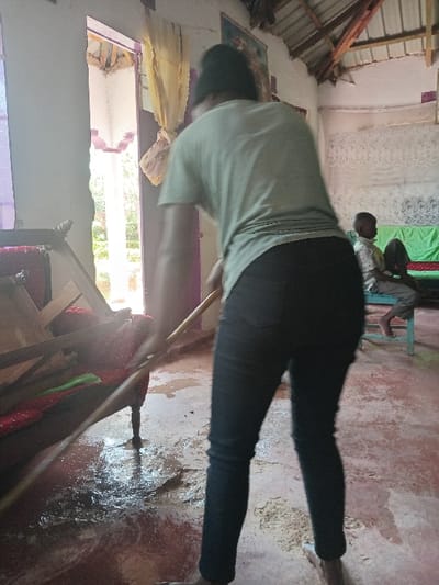This image depicts an interior scene within a structure located in Manda-Shivanga ward, Kenya. In the foreground, an individual, seen from the rear, is pushing water and dark debris across a reddish-brown concrete floor using a long wooden pole or broom handle. This individual is barefoot, wears dark trousers, a light-colored short-sleeved shirt, and a dark head covering. The floor is extensively wet with standing water, mud, and debris, indicative of a flood cleanup operation. To the left of the active cleanup, a wooden daybed or bench with red and green cushioning is partially submerged in the water and debris. In the background, a male individual, appearing to be a child or young adult, is seated on a light blue stool, facing towards the right side of the frame. He wears a light-colored patterned shirt. Behind him, a surface is covered with green fabric. The room features white-painted walls, with an open doorway visible on the left, revealing an outdoor area with vegetation. The ceiling consists of exposed wooden rafters and corrugated metal sheeting, with white fabric elements suspended in the upper right portion of the frame. A framed picture hangs on the wall in the upper left background.