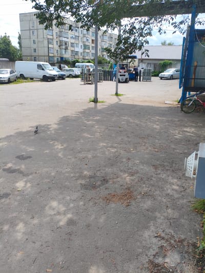The image depicts an outdoor scene with an apartment building in the background. Several vehicles, including white vans, a car, and a forklift, are parked in a lot. There are also gas cylinders stacked and being moved by a forklift. People are visible interacting with the equipment and vehicles. A pigeon is walking on the paved ground in the foreground. A bicycle is parked on the right side of the image. The overall environment suggests an industrial or storage area within the city of Lubny, Ukraine.
