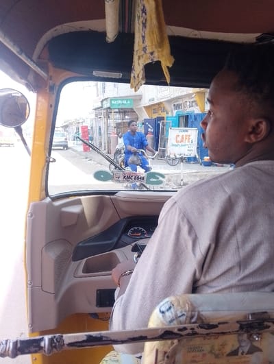 The image captures a street scene in Dorayi, Nigeria, from the interior of a yellow motorized tricycle. In the foreground, the right shoulder and arm of a person, likely the driver, wearing a light-colored garment, are visible. Their right hand rests on the steering mechanism, and a dashboard is present. A rearview mirror is affixed to the left, and a patterned yellow fabric hangs from the tricycle's roof.
Through the windshield, a young man on a bicycle, dressed in a blue traditional outfit, rides across the midground from right to left. The bicycle features a white sign attached to its front displaying "SKATUR KANYA" and "KMC 8848". The street appears paved. On the right side of the street, multiple gas cylinders are visible outside a storefront.
In the background, a row of commercial buildings lines the street, featuring various signs. Identifiable signs include "NASHI OIL" with green text, "GURU GAS", "TWINS ELECTRICA", and a prominent white sign for "SANCHI CAFE & Restaurant". Another vehicle is partially visible further down the street on the left. The scene is illuminated by daylight.