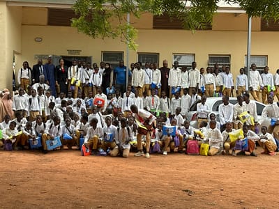 This outdoor image captures a large gathering of predominantly young male students and several adults in Unguwan Dosa, Nigeria. The scene is set in front of a single-story, light-colored building, likely a school hall, with an unpaved reddish-brown dirt ground. Green foliage from an overhead tree provides shade at the top of the frame, and parts of white vehicles are visible behind some of the students on the right.

The majority of the subjects are students, dressed uniformly in white long-sleeved shirts and khaki or tan trousers. Many are either standing, kneeling, or crouching in the foreground and mid-ground. They are holding various colorful items, including small purple, yellow, and red bags, as well as blue folders. Several of the blue folders are clearly visible with text that reads "NEEC Essential" and a corresponding logo. Some students also have backpacks.

A group of adults stands behind the students. These adults include men dressed in dark suits, traditional Nigerian attire (long robes and caps), and casual wear like a blue polo shirt. Women are also present, dressed in formal wear, including suits. These adults appear to be organizers or benefactors, participating in what seems to be a distribution event, likely of school supplies.

Notable details include a sign on the building stating: "THIS HALL WAS RENOVATED IN AND OUT NOV. 2021 BY SAMORA GISEI." Another, smaller, less legible sign is to the left of the main door. The event takes place during the daytime, indicated by the bright, even lighting. The atmosphere appears orderly, with some students showing smiles, suggesting a positive and organized activity.