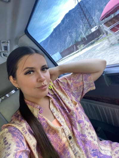 A young woman with dark hair pulled back in a ponytail is seated in the passenger seat of a vehicle, her right arm resting behind her head. She is wearing a patterned shirt with shades of purple, orange, and cream, and an orange camisole is visible underneath. Her nails are painted a neon yellow-green. The interior of the car is dimly lit, with a glimpse of a grey leather seat. Through the rear window, a mountainous landscape under a cloudy sky is visible. The exterior scene shows a dirt or gravel road, a fence, some structures, and a tall metal pylon. The overall impression is a casual, candid shot taken during a road trip in a scenic, mountainous region. The location is identified as Irganai, Russia.