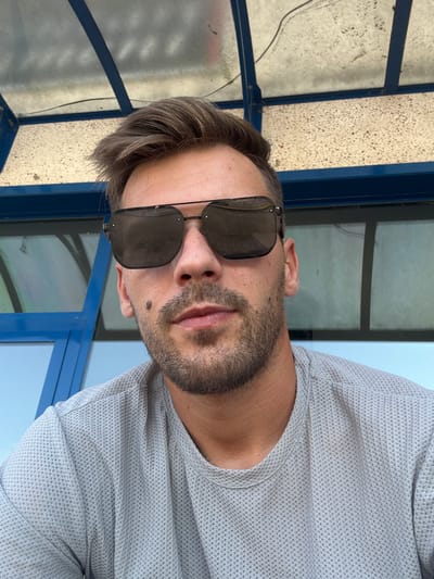 A close-up, outdoor, selfie-style portrait shows a man in his late 20s or early 30s wearing sunglasses and a light gray, waffle-knit t-shirt. He has neatly styled brown hair, a short beard, and a mole on his left cheekbone. The man is positioned in the foreground, with a blue and white striped awning and blue framed windows visible in the background. The setting appears to be a public area with a backdrop of windows and a partial view of an awning structure, suggesting an urban or semi-urban environment possibly near a sporting facility or park. The lighting is natural, indicating it is daytime. The overall impression is casual and relaxed. The provided context places the scene in Castelletto sopra Ticino, Italy.