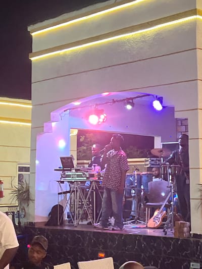 In the city of Oba, Nigeria, a band performs on a stage at night. The stage is lit with colorful lights, casting a vibrant glow on the performers. A lead singer, wearing a checkered shirt and jeans, stands center stage with a microphone. To his right, a drummer is seated behind a drum kit, and another musician is visible behind a DJ setup. A guitar rests on a stand to the right of the stage. The stage is elevated and appears to be outdoors, with a white arched facade featuring yellow LED strip lighting overhead. In the foreground, audience members are seated, with one person in a cap visible. The overall atmosphere suggests a lively musical performance.