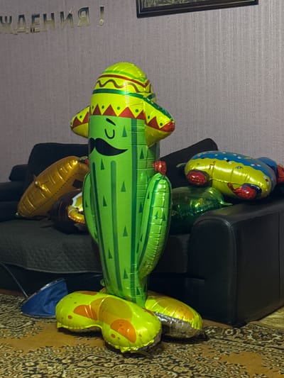 A novelty cactus-shaped balloon with a sombrero and mustache is the main subject in a dimly lit room, possibly a living room. The balloon is green and stands upright, with its base resting on a patterned rug. Other balloons are visible, including a gold, elongated balloon to the left and a yellow and blue balloon shaped like a creature with wheels to the right, both resting on a dark sofa. Behind the balloons, a textured wall is visible with golden lettering that spells out "ЖДЕНИЯ!". A dark sofa is prominent in the mid-ground, and a portion of a dark armchair is visible on the right. The floor is covered with a detailed, ornate rug. The lighting in the room appears to be artificial and casts shadows. The location is stated to be Фёдорово, Russia, though this context is not visually evident from the image itself.