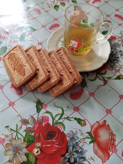 A tabletop scene displays a glass mug filled with amber-colored liquid, likely tea, adorned with cartoon characters. Beside the mug, on a white saucer, are several rectangular biscuits, each imprinted with a corn cob design. The tabletop itself is covered with a patterned cloth featuring a grid of red lines and pink diamond shapes, interspersed with painted floral designs, including red roses and daisies. The lighting suggests an indoor setting. This scene is located in Luka, Ukraine.