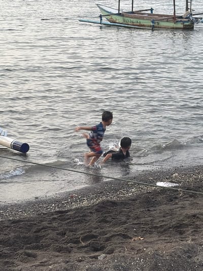 The image depicts a scene at a beach in Buleleng, Indonesia. Two young children are playing in the shallow water near the shore. One child, wearing a black t-shirt, is partially submerged in the water, with water splashing around them. The other child, wearing a colorful patterned shirt and shorts, is in a crouching position, also interacting with the water.

The water has a rippled texture, suggesting gentle movement. To the left of the frame, a traditional Indonesian fishing boat, painted green with the name "MANDALIKA" visible in faded lettering, is moored near the shore. The boat has outriggers and appears to be resting on the water.

The shoreline is composed of dark, coarse sand mixed with small pebbles. A dark rope runs diagonally across the image, from the upper right towards the lower center, suggesting a mooring line or boundary. Part of a white object with a blue tip, possibly a buoy or part of another boat, is visible in the lower center.

The lighting suggests it could be daytime, possibly late afternoon given the angle of light on the water, but it's difficult to determine precisely. The overall mood appears to be one of carefree childhood play in a natural coastal environment.