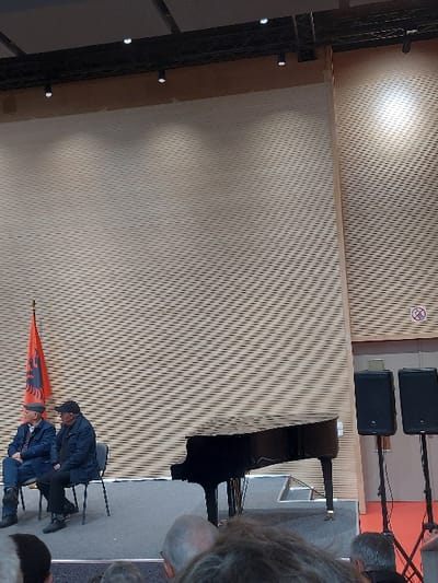 This indoor image captures a formal event, likely a concert or ceremony, in Tiranë, Albania. The scene features a stage or elevated platform with a grand black piano positioned prominently on the right side. Above the piano, two black speakers are mounted on stands. A circular white sign with a red border and a diagonal line, indicating "No smoking," is visible on the wall directly above the speakers.
The background is dominated by a light-colored wall with a textured, patterned design composed of diagonal lines forming small diamond shapes, possibly acoustic paneling. Bright spotlights illuminate sections of this wall, creating distinct highlights.
In the foreground, an Albanian flag, displaying its characteristic red field with a black double-headed eagle, stands upright on the left. Two men are seated on chairs near the flag. One man, wearing a dark jacket and a cap, is partially obscured by the flag. Next to him, another man, dressed in a blue jacket and a cap, is also seated. The blurred heads and shoulders of several other people are visible in the immediate foreground on the right, suggesting an audience or attendees for the event. The setting implies a structured gathering or performance.