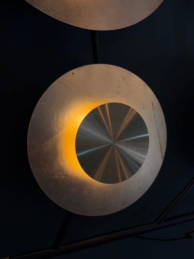 A close-up shot features a modern decorative element, likely part of a lamp or art installation, set against a dark, uniform background. The central subject is a circular object composed of two concentric rings.

The outer, larger ring has a matte, textured metallic finish, possibly silver leaf or a brushed, lightly distressed metal, exhibiting subtle variations in tone and some visible scratches. A warm, golden light emanates from the lower inner edge of this outer ring, casting a soft glow onto its surface and the inner circle.

The inner, smaller circle is made of a highly reflective, brushed metallic material, such as stainless steel or aluminum. It features distinct radial lines originating from its center, which create a starburst or sunburst effect as they catch and reflect the light. This central reflective part bounces the warm light from below, creating a captivating interplay of light and shadow.

To the left, a portion of a similar circular element is visible, echoing the design and material of the main subject. On the right, a dark, angular structure, likely part of the fixture's frame or stand, is partially visible, providing a dark contrast. The overall impression is one of sleek, contemporary design with a warm, inviting glow, suggesting an indoor setting designed for ambient lighting. No people or visible text are present. The location context provided is Dobrota, Montenegro.