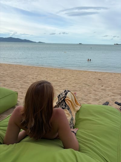 In Ko Samui District, Thailand, a woman with brown hair is reclining on a large, lime green beanbag cushion on a sandy beach. She is facing away from the camera, towards the ocean. She wears a yellow and black patterned sarong draped over her lower body. Her right arm is bent, resting on the cushion, and she appears to be wearing a bracelet on her left wrist. Her feet, one with pink nail polish, are visible near the bottom of the frame. In the background, the calm ocean stretches to the horizon, with a distant, mountainous island to the left and smaller islands or landmasses further out. Two people are visible swimming in the sea. The sky above is cloudy with patches of blue. The beach sand is light brown, and another green cushion is partially visible to the left. A pair of black flip-flops rests on the sand to the right of the beanbag.