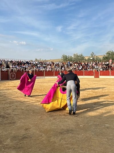 The image shows a bullfighting scene in the city of San José del Valle, Spain. In the center of a sandy bullring, two matadors are visible, one holding a pink cape while facing the bull, and another wearing grey pants and a dark coat standing near a yellow and pink cape on the ground, seemingly taunting the bull. In the background, a crowd of spectators is seated in the stands. The sky above is blue with scattered clouds.