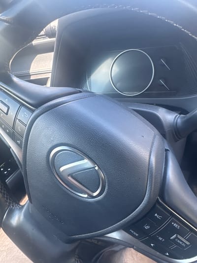 The image shows a close-up view of the interior of a car, focusing on the steering wheel and dashboard. The steering wheel is black leather with a chrome Lexus logo in the center. To the left of the steering wheel, there are buttons for media and phone controls. To the right, there are buttons for cruise control and mode selection. The dashboard features a digital display screen and two analog gauges. The lighting suggests it is daytime, with sunlight coming from the upper left, casting shadows on the steering wheel. The overall condition of the interior appears to be clean. There are no people visible in the image. The location is not specified in the image itself, and therefore cannot be determined to be in Unknown, Canada or any other location.