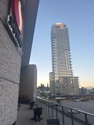 The image shows a Safeway grocery store in Coquitlam, Canada, as seen from an elevated perspective. The foreground features the exterior of the Safeway building, with its distinctive red and white logo prominently displayed. Adjacent to the Safeway, there is an outdoor seating area with tables and chairs. In the background, a tall, modern high-rise building dominates the skyline, with its glass facade reflecting the clear blue sky. Below the high-rise, there are various commercial establishments, including a Dollarama store, and a parking lot with several cars. Residential buildings and trees can be seen further in the distance, suggesting a mixed urban and suburban environment. The time of day appears to be late afternoon or early evening, judging by the soft lighting and clear sky.
