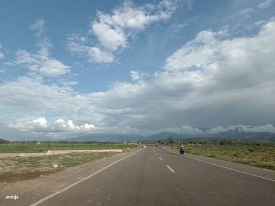 A wide, asphalt road, marked with a white solid line on the left and dashed white lines in the center, curves gently to the right, disappearing into the distance. The road is flanked by dry, grassy terrain on both sides, with some scattered vegetation. In the far distance, a range of low, green mountains are visible, partially obscured by a dramatic sky filled with a mixture of white, puffy clouds and dark, heavy storm clouds. On the right side of the road, a motorcycle with a rider wearing a helmet is in motion. In the distance on the left, a flat, green landscape, possibly agricultural fields, stretches out. A few structures, including what appear to be construction materials and small buildings, are scattered within this landscape. The overall impression is that of a rural, open-air environment under a dynamic sky. The location is identified as Dayah Kruet, Indonesia.