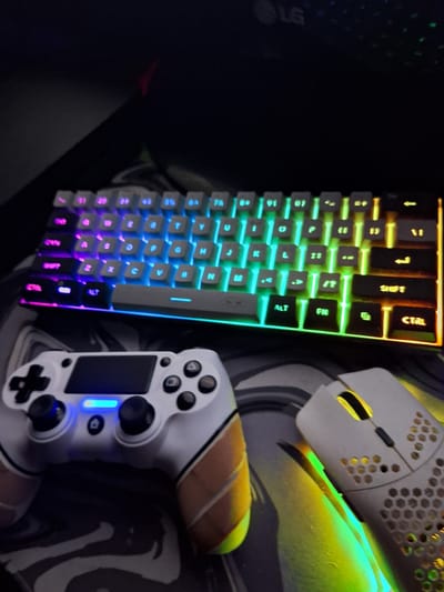 The scene is located in Kornitsa, Bulgaria. A dark, low-light environment displays a gaming peripheral setup. A compact mechanical keyboard with RGB backlighting illuminates in a gradient from purple on the left to blue, green, and orange on the right. Below and to the left of the keyboard, a white PlayStation 4 controller is positioned, featuring custom brown adhesive grip tape on both handles and an illuminated blue light bar. Below and to the right of the keyboard, a white gaming mouse with a honeycomb perforated shell exhibits an internal yellow-orange light. All three peripherals rest on a black mouse pad featuring a gray and white abstract pattern. In the upper background, a dark monitor with an "LG" logo is partially visible.