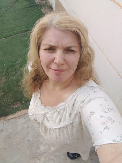 A medium shot, selfie-style photograph taken outdoors shows a fair-skinned woman with blonde hair, likely in her 40s or 50s, looking directly at the camera. She is wearing a light-colored, floral-patterned top with lace trim and a small bow at the neckline. Her expression is neutral to slightly strained, with her brow furrowed. Her skin shows some redness and visible pores.
The woman is positioned in what appears to be a residential or balcony area. To her left, a patch of green grass, possibly a lawn or field, is visible, demarcated by a subtle curve in the ground. The ground beneath her feet is a textured, light gray concrete surface. Behind her, to the right, is a section of a building wall, characterized by a pale, off-white color and faint vertical lines or seams. The overall lighting suggests it is daytime, with a slightly overcast or diffused light source. The image quality is somewhat grainy. No specific landmarks or indicators of the city of Al Mas'oudi, United Arab Emirates, are discernible within the frame.