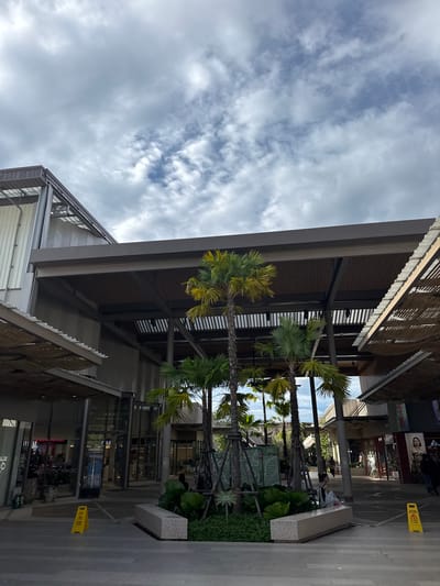 The image captures an outdoor scene at what appears to be a shopping center or mall in Krabi, Thailand. The perspective is from a low angle, looking upwards towards a partially covered walkway or building. Palm trees with lush green fronds are prominently featured, with some reaching towards a sky filled with dynamic clouds. The weather appears to be partly cloudy, with patches of blue sky visible amidst the white and grey clouds.
The architecture of the building features modern design elements, including metal beams, a dark-colored facade, and large glass windows that reveal glimpses of interior spaces. One of the glass doors has the word "FREE" visible on it, possibly indicating a promotion or service. In the foreground, a concrete bench is situated next to a planter box filled with dense green foliage. Further down the walkway, a yellow caution sign is propped up, indicating a wet floor.
Several people are visible in the background, walking along the shaded pathway. Their presence suggests that the location is open and accessible. The overall mood is one of a bright, possibly warm day, with a sense of openness and a tropical ambiance due to the presence of the palm trees. The time of day seems to be daytime, with the lighting suggesting it is not yet dark. The scene evokes a sense of leisurely activity, perhaps shopping or strolling.