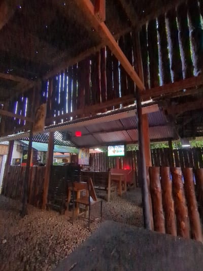 The image shows the interior of a wooden-framed building in Morogoro, Tanzania, located at coordinates -6.835 latitude and 37.653 longitude. The structure appears to be a bar or restaurant, featuring a wooden slatted ceiling with a single light bulb hanging from it, and wooden walls and beams. An obscured television screen is visible mounted on the wall, displaying a sporting event. Several wooden tables and chairs are arranged in the space, which has a gravel floor. One wooden bench sits against a wall, with other chairs and tables scattered around the open area. The building is dimly lit and has a rustic feel.