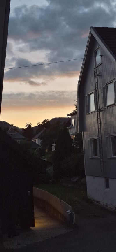 An outdoor, vertical, full-frame, medium-shot photograph shows a residential area with houses nestled against a hillside under a cloudy twilight sky. In the foreground, to the left, is the dark side of a building. To the right, a grey, multi-story house with a steeply pitched roof dominates the frame. This house has several windows, some with dark shutters, and a metal fire escape ladder running vertically up its side. In the middle ground, several other houses are visible, appearing partially obscured by trees and the darkening light. The sky above displays a gradient of colors, from darker grey clouds at the top to softer, warmer hues of orange and pink closer to the horizon, suggesting the end of the day. The environment appears to be a hillside community, likely in Lillehammer, Norway, given the architecture and terrain which are consistent with the area. There are no people or animals visible in the image. The overall scene conveys a sense of quiet dusk in a rural or semi-rural residential setting.