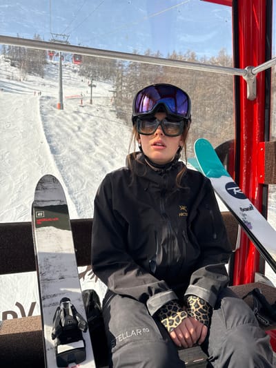 The image captures a female individual seated within a red-framed gondola cabin at a ski resort in Saint-Chaffrey, France, under a clear blue sky. The individual wears a black helmet, with reflective purple ski goggles pushed up, and dark-framed sunglasses over her eyes. She is dressed in a black Arc'teryx ski jacket, black ski pants displaying "STELLAR EQ" on the left thigh, and leopard-print wrist gaiters. Her expression is neutral with her mouth slightly open. To her left, an ARMADA WHITWALKER 116 ski with black bindings rests upright against the dark brown seat. To her right, a white Rossignol ski with blue-green tips and black graphics is similarly positioned. Through the gondola window, a snow-covered ski slope descends, revealing a multi-pylon ski lift system with several red cabins and scattered evergreen trees in the mid-ground. Orange boundary markers are visible on the distant slope.