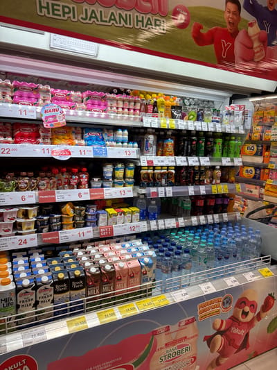 This image captures a brightly lit refrigerated display aisle within a supermarket or convenience store, likely in Kelapa Dua, Indonesia, given the currency and product labels. The perspective is tilted, showcasing a wide array of beverages.

The primary subjects are rows of bottled and cartoned drinks. On the far left, multiple packs of Yakult fermented milk drinks are prominently displayed, including "STROBERI" (Strawberry) and "MANGGA" (Mango) flavors. A large sign for "Yakult" features a man in a red jersey with a 'Y' logo, excitedly holding a bottle, accompanied by text like "HEPI JALANI HARI" and "VITAMIN Y". Another large promotional graphic on the right advertises "Yakult RASA STROBERI" with a superhero-like character, emphasizing "PROBIOTIK SETIAP HARI".

Further into the display, various other products are visible:
*   **Water bottles:** Rows of bottled water (e.g., Purified) and some soft drinks like Sprite.
*   **Milk:** Numerous milk cartons including "Cimory 100% FRESH MILK", "Brook Farm Fresh Milk", "Diamond Fresh Milk", "Milk Life lactose free milk", and flavored milk (chocolate, strawberry).
*   **Yogurt and other drinks:** Products like "Skim Milk", Sunkist juice, "Fibe" drinks, Golda Coffee, Kin drinks, and various yogurts such as "Yoseo", "Dezzert Milky", "Biokul", and "Heavenly Blush Greek".

Each product section has clear price tags displaying prices in Indonesian Rupiah (Rp), with several promotional offers like "PROMOTION", "BUY 2 GET 1", "PRODUK BARU" (New Product), and "GRATIS Voucher Rp50rb" (Free Rp50rb Voucher). The prices range significantly, for example, Yakult at Rp 11.800/pack, Diamond Fresh Milk at Rp 24.500, and Brook Farm Fresh at Rp 33.500. Some background shelves show packaged goods like "nic" and "TISS" tissues. The "HALAL" logo is visible on some Yakult products, affirming their suitability for Muslim consumers.
