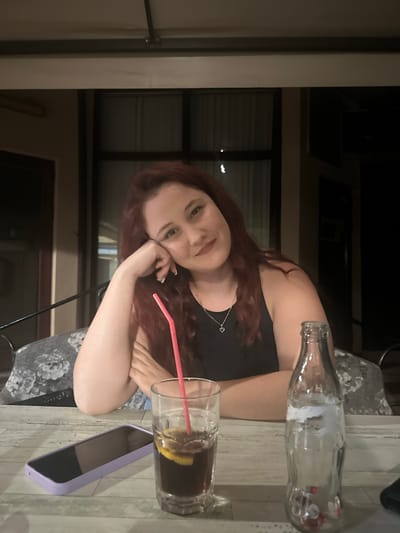 A young woman with auburn hair, wearing a black tank top and a silver necklace with a heart pendant, is seated at a table outdoors. She is resting her head on her hand, looking at the camera with a slight smile. On the table in front of her are a glass of dark liquid with a pink straw and a slice of lemon, a light purple smartphone, and a clear glass bottle with some residue inside. The background shows the exterior of a building with dark windows and a covered patio area. The scene suggests a relaxed evening at an outdoor cafe or restaurant in the city of Lom, Bulgaria.