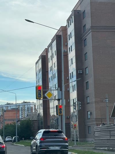 This image captures a street scene in the city of Petropavl, Kazakhstan. A modern, multi-story brick building with large, reflective windows dominates the right side of the frame. In front of the building, a traffic light displays a red signal, and a diamond-shaped yellow warning sign is visible. Further down the street, another traffic light shows a red signal. Several cars are on the road, including a dark-colored SUV in the foreground with a license plate that reads "975 ADT 15". In the background, a low-rise building with a gray, sloped roof is situated between the modern building and other residential structures. The sky is partly cloudy, with patches of blue.
