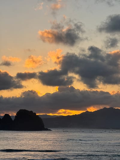 The image captures a dramatic sunset over the ocean at Selongbelanak, Indonesia. Dark, textured clouds are illuminated from behind by the setting sun, creating a fiery orange and yellow glow along their edges. These vibrant clouds contrast sharply with the darker, more opaque clouds in the foreground.

In the distance, the silhouette of a dark, possibly forested, headland juts out into the ocean. The sea itself is calm, with gentle, reflective ripples on its surface, catching the muted light of the twilight sky. A thin line of what appears to be land or a distant island can be seen on the horizon, almost blending with the sky. The overall mood is serene and awe-inspiring, emphasizing the natural beauty of the sunset. The time of day is clearly evening, as indicated by the sunset. There are no visible people, objects, or text in the image.