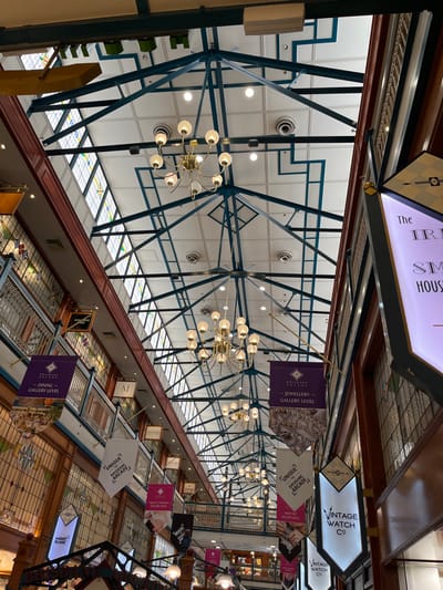 This media file captures an overhead view looking down a multi-level indoor shopping arcade, likely the Brisbane Arcade in Brisbane City, Australia. The scene is brightly lit, suggesting daytime, with natural light filtering through extensive skylight panels integrated into a high, vaulted ceiling.

The ceiling features an intricate geometric framework of dark blue or black metal beams, creating a striking visual pattern. Numerous ornate brass chandeliers with frosted globe bulbs hang at regular intervals, providing artificial illumination alongside recessed circular lights in the ceiling panels.

A dark wooden railing on the upper level, where the photo is taken from, offers a clear view of the levels below. These lower levels feature ceilings or walkways adorned with decorative stained glass patterns.

Suspended throughout the arcade are various informational and directional banners and signs. Visible text includes:
*   "VINTAGE WATCH CO"
*   "UNIQUE BRISBANE ARCADE" (reinforcing the location cue)
*   "-JEWELLERY GALLERY LEVEL"
*   "-DINING- GALLERY LEVEL"
*   "MONTO BLANC" (partially visible)
*   "MADE"
*   "M" (a logo)
*   "The IR SM HOUS" (partially visible)

The overall impression is that of a well-maintained, possibly historic, upscale commercial space, likely housing various retail and dining establishments. No people are visible, but the setting implies a commercial environment open for business.